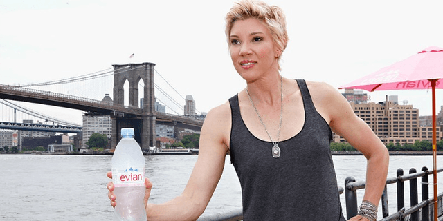 Jackie Warner holds an Evian bottle while posing in a tank top in front of a bridge and a river