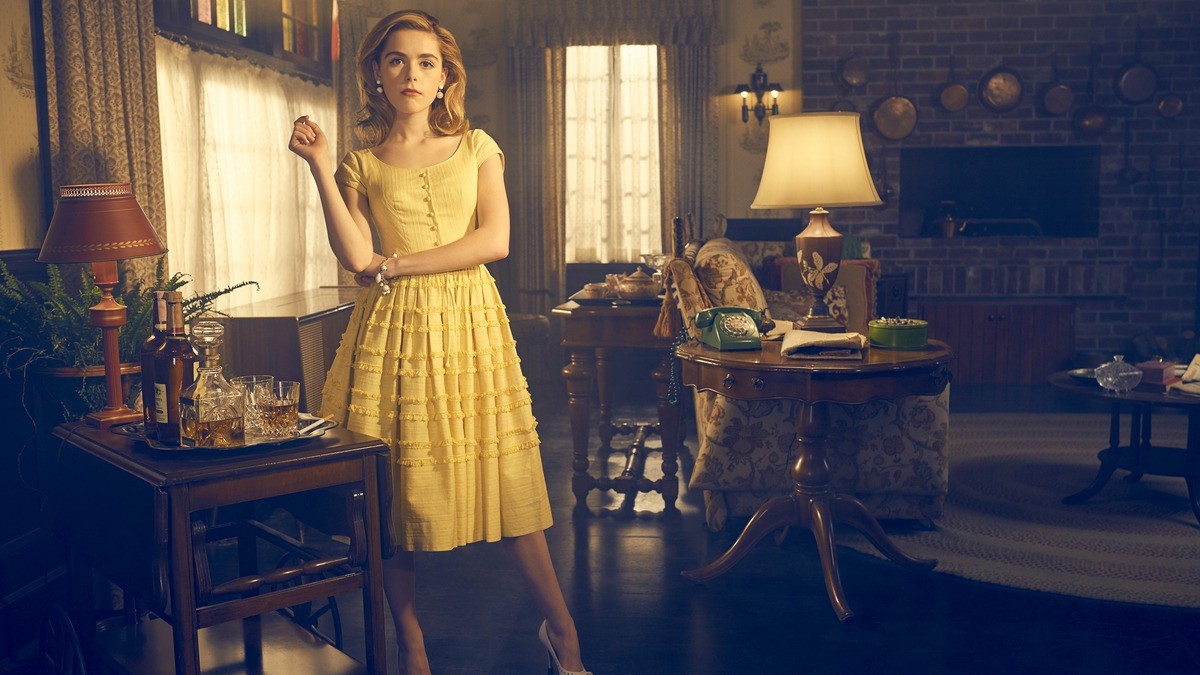 B.D. Hyman stands in an front of a minibar in an old-fashioned living room, wearing a yellow dress, in a promotional image from the FX series 'Feud.'