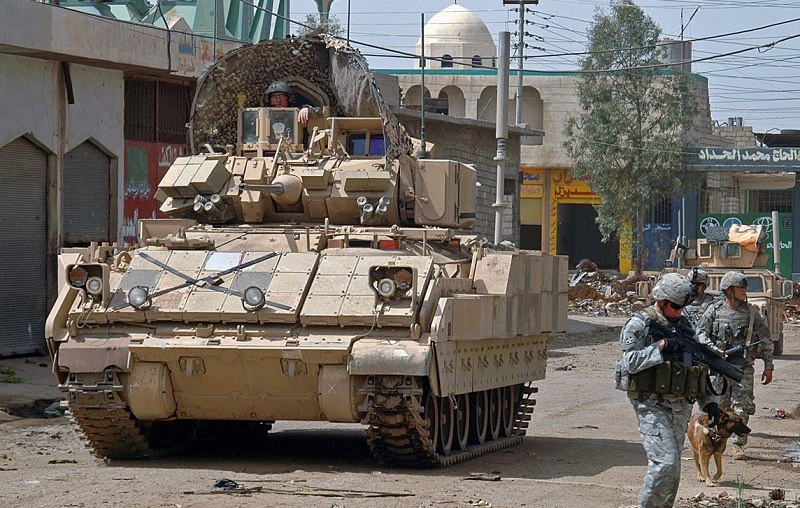 An M2 Bradley Fighting Vehicle rolls down a street while soldiers walk nearby.