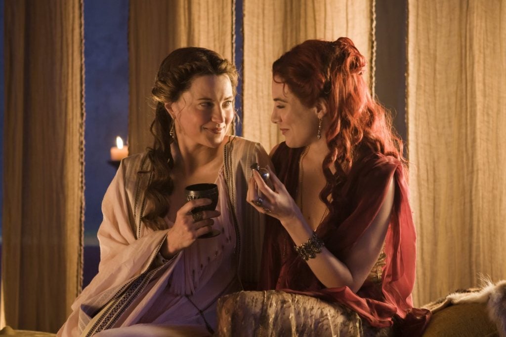 Lucy Lawless and a woman smile together on a bed in Spartacus