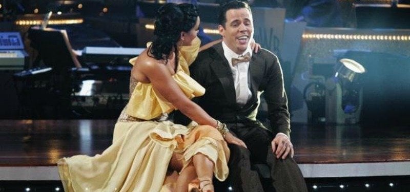 Steve-O is sitting on the steps on stage as Lacey Schwimmer has an arm around him and is looking at him.