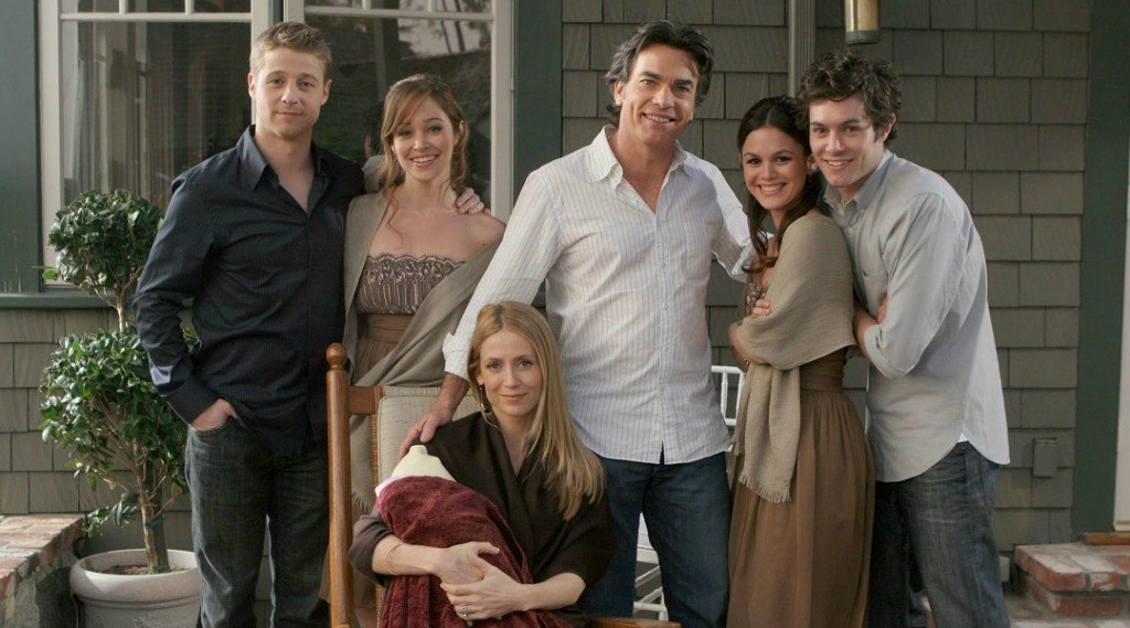 Ben McKenzie as Ryan Atwood, Autumn Reeser as Taylor Townsend, Kelly Rowan as Kirsten Cohen, Peter Gallagher as Sandy Cohen, Rachel Bilson as Summer Roberts, and Adam Brody as Seth Cohen with their arms around each other in front of a house on The O.C. 