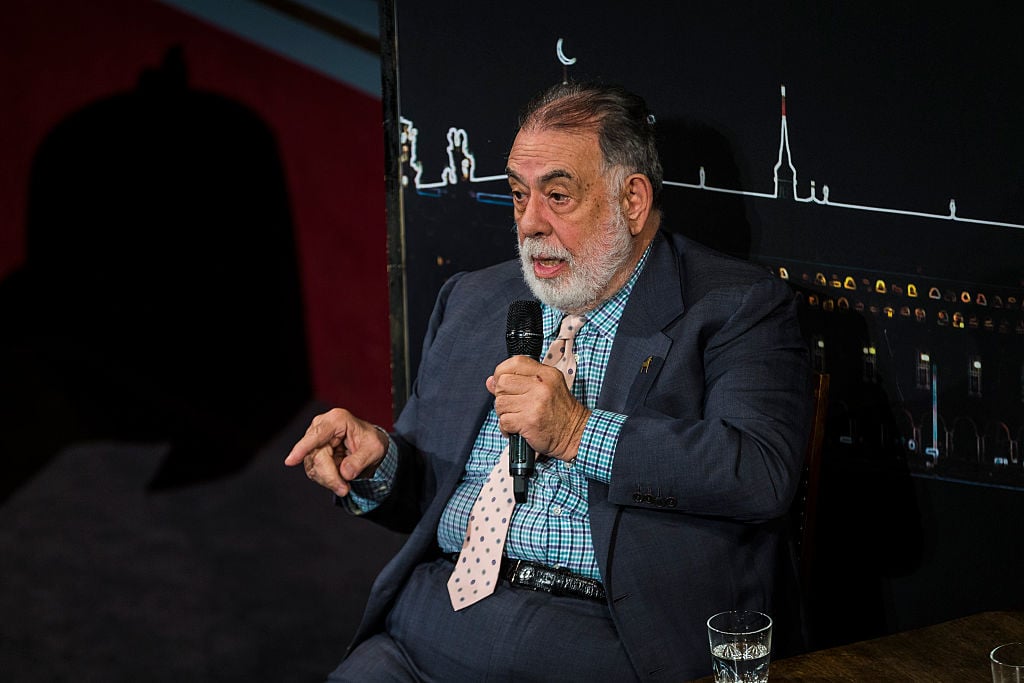 Francis Ford Coppola, sitting in a chair, speaking into a microphone, and pointing with his right hand