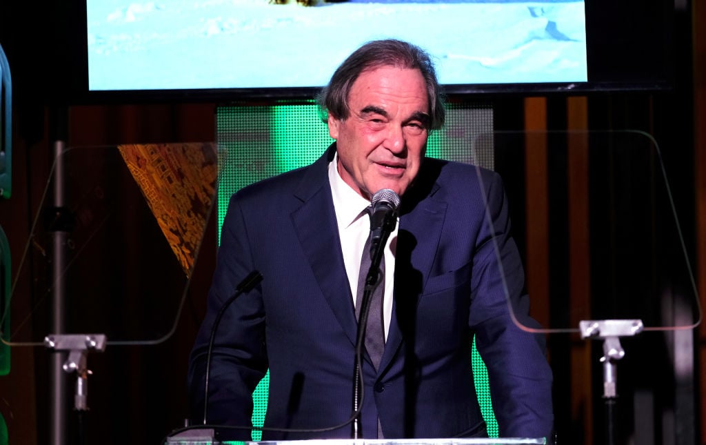 Oliver Stone in a suit, speaking at a podium