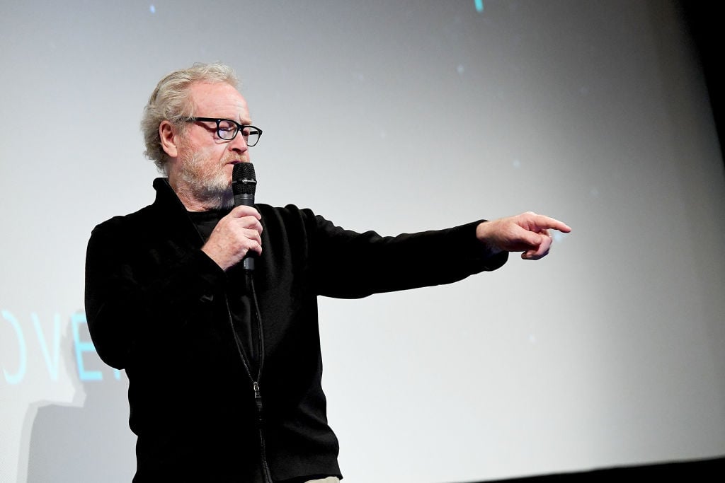 Ridley Scott wearing all black, speaking into a microphone, and pointing with his left hand