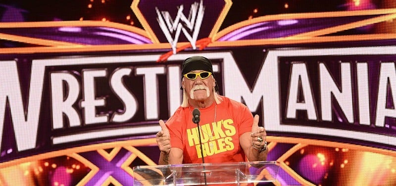 Hulk Hogan is giving two thumbs up at a podium in front of a Wrestlemania poster.