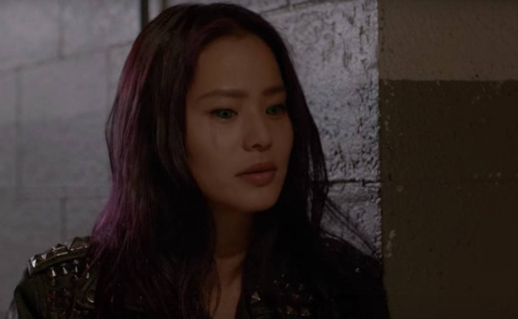 Jamie Chung with purple hair and eyes, leaning against a wall and looking downward