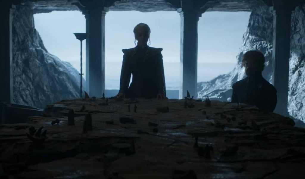 Daenerys stands over a table shaped like Westeros, with Tyrion off to her left