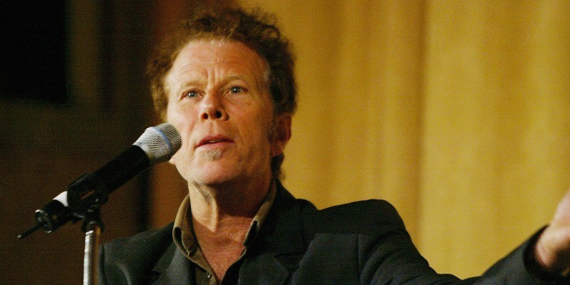 Tom Waits stands in front of a microphone and holds his arm out.