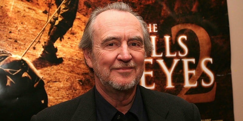 Wes Craven stands in front of a 'The Hills Have Eyes 2' poster.