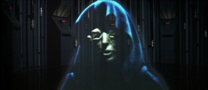 A hologram of Emperor Palpatine, shrouded in shadow and wearing a black hood