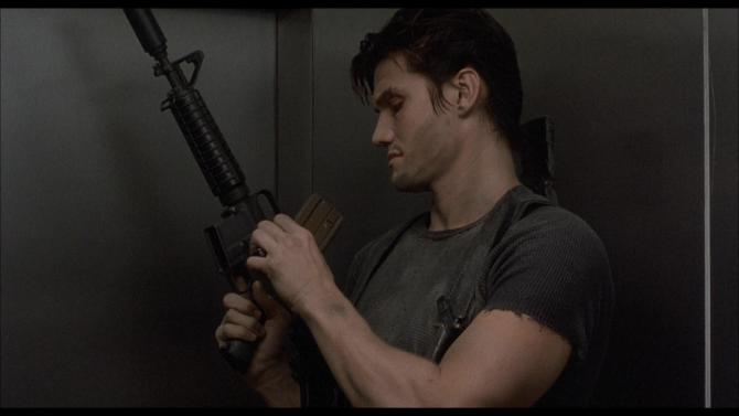 Dolph Lundgren, loading a large gun and wearing a grey tshirt