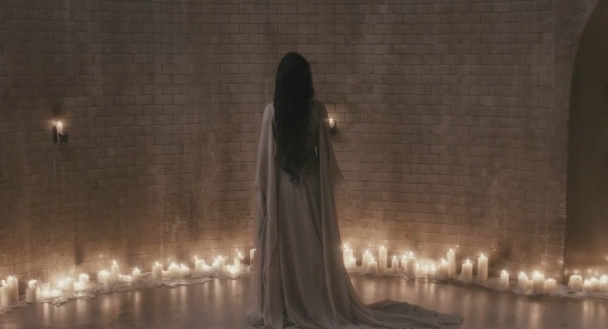 Eva Green in a white robe facing away in front of a brick wall and many candles