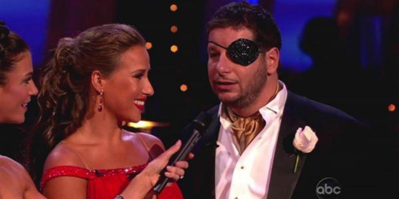 Jeffrey Ross is wearing an eye patch and is talking into a microphone as Edyta Sliwinskaare looks at him smiling.