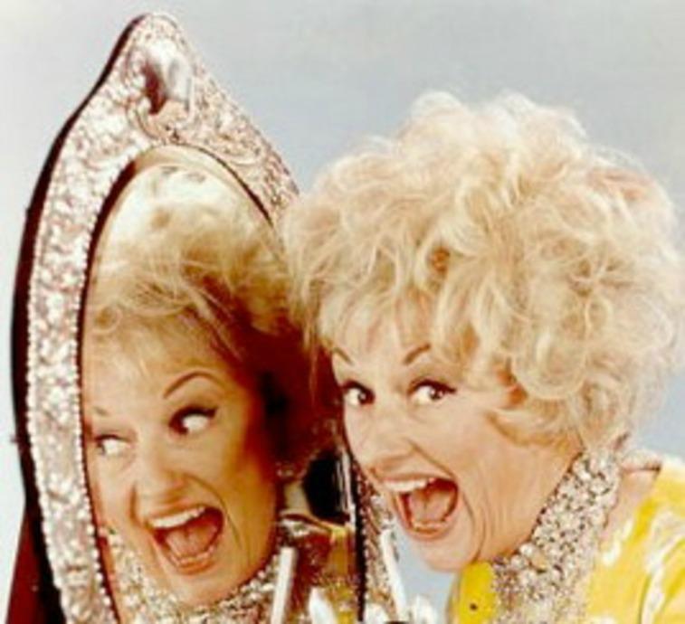 Phyllis Diller smiling with her mouth open wide next to a mirror where you can see her reflection
