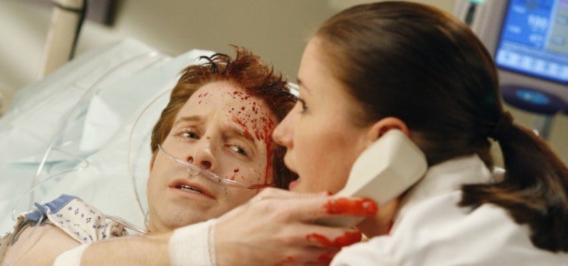 Seth Green is covered in blood in a hospital bed and is holding up a phone to a doctor's ear.