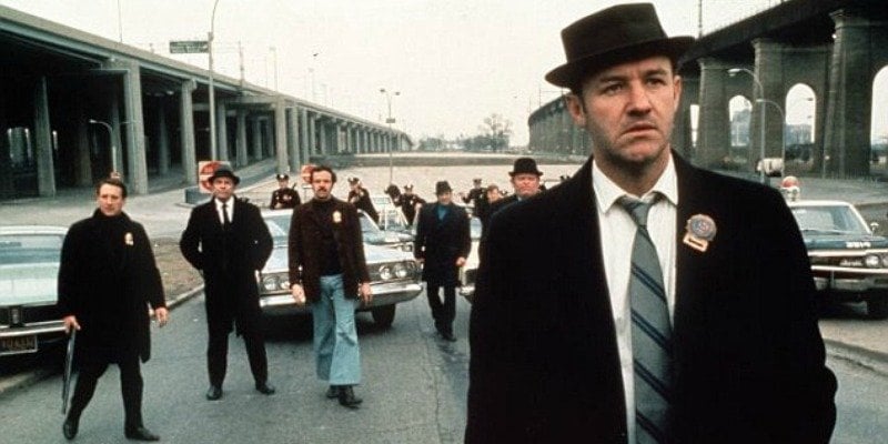 Gene Hackman is in a suit and is wearing a hat. He is standing in front of a group of men in The French Connection.