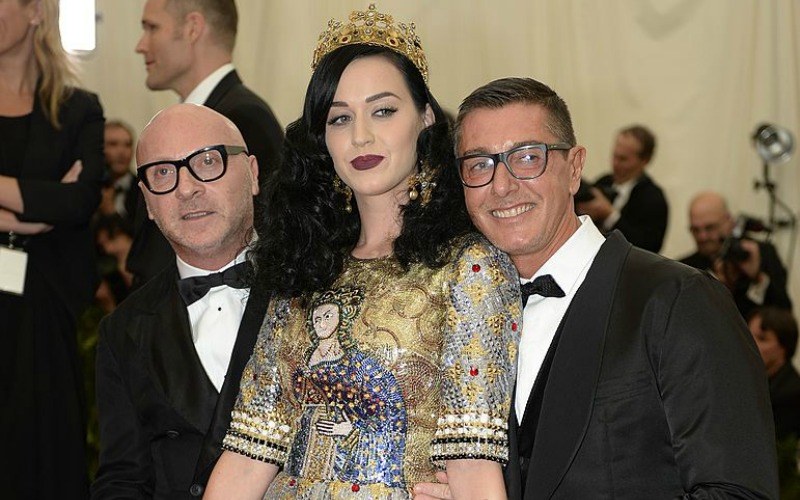 Katy Perry is in a gold dress and is wearing a crown with two men in tuxedos beside her.