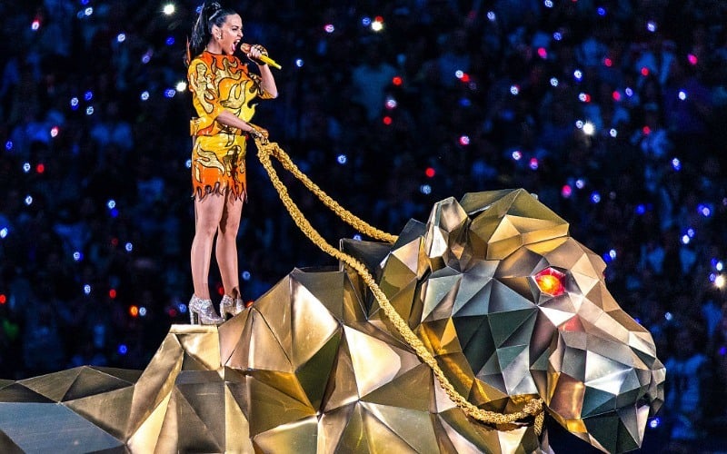 Katy Perry is in a shirt and shorts that are made to look like fire while holding the reigns to a metallic animal.