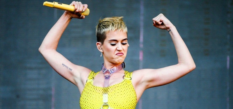 Katy Perry flexing her muscles in a yellow top holding a yellow microphone
