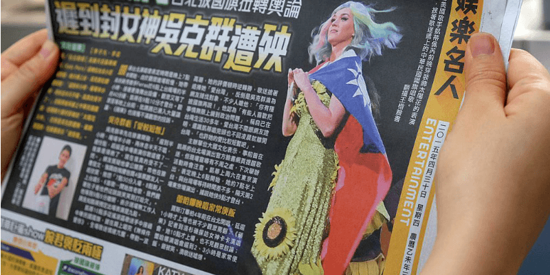 A hand holding a Chinese newspaper which shows Katy Perry wearing a sunflower dress and using a flag as a cape.