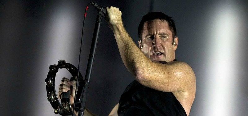 Trent Reznor is holding a microphone in one hand and shaking a tambourine with the other on stage.