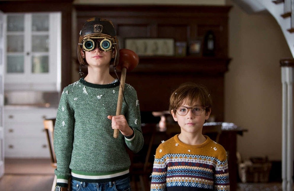 Two children stand facing a camera, with one holding a plunger over his shoulder and wearing goggles
