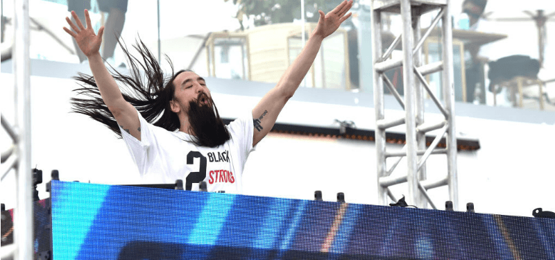 Steve Aoki has his hands in the air behind a DJ booth.
