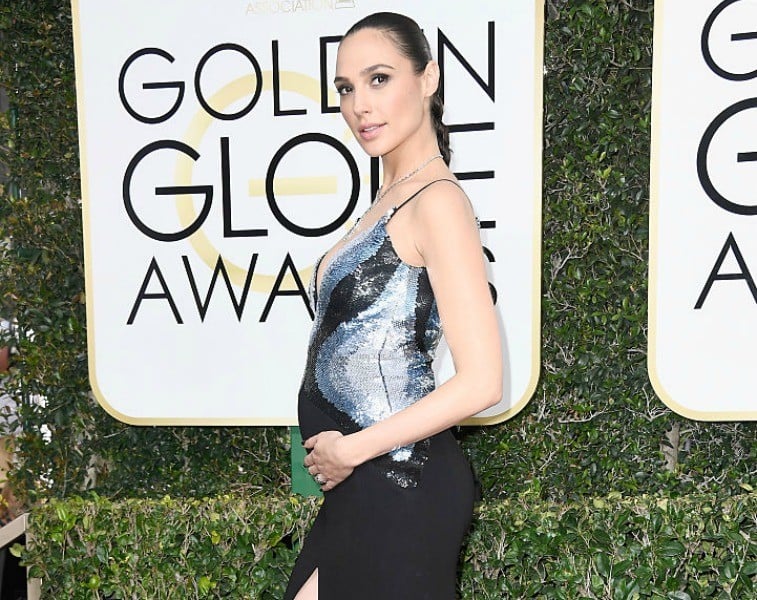 Gal Gadot poses in a black and silver dress with her hand on her belly in front of a sign for the Golden Globe Awards