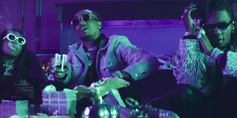 Members of group Migos are sitting down on a couch smoking and drinking in a club