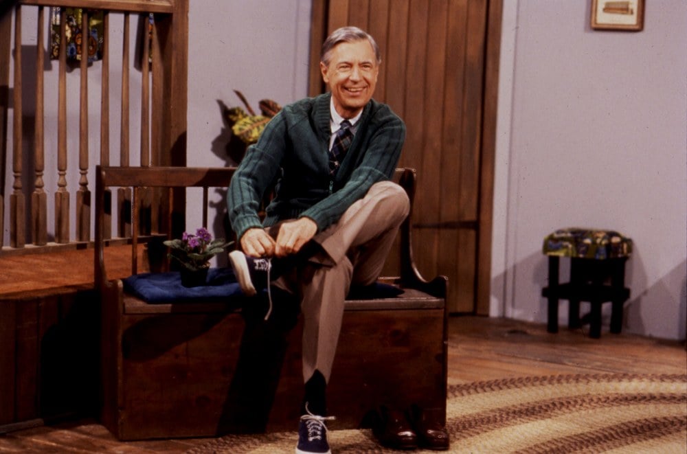 Fred Rogers sits on a bench on set taking off his shoes