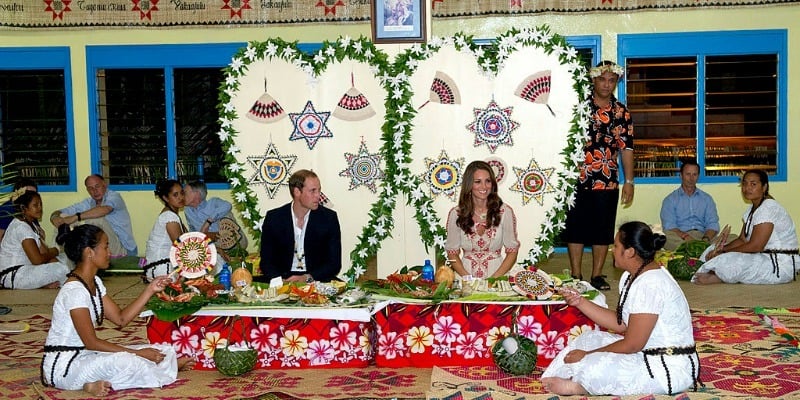 Kate Middleton and Prince William are sitting down in huge chairs at a dinner table as two woman sit down on the ground next to them.