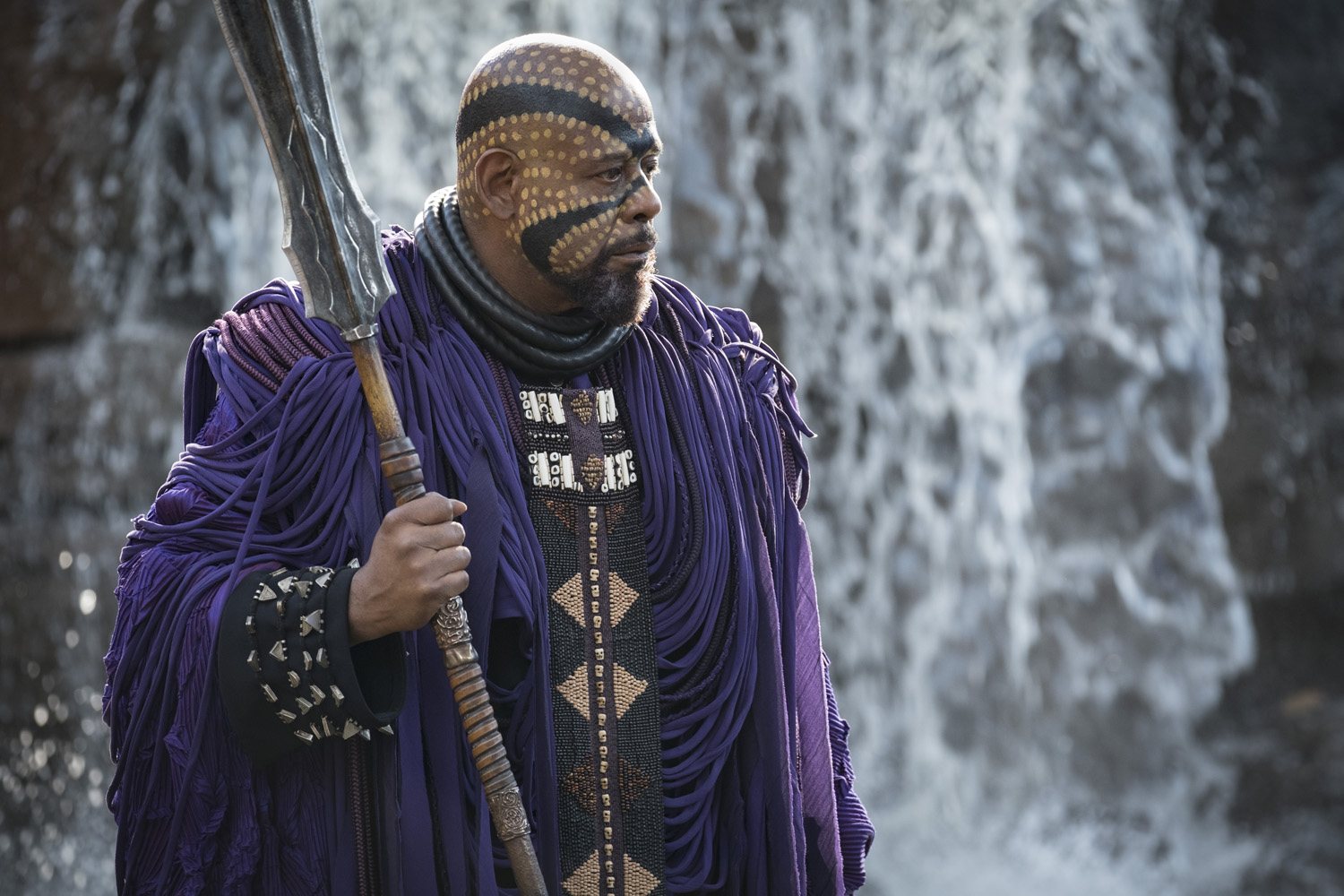 Forest Whitaker's Zuri wears purple garb and elaborate face paint while holding a giant scepter in Black Panther