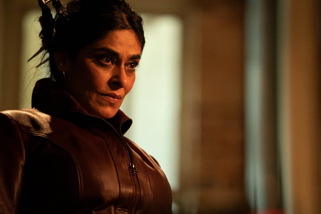 Nimra Bucha als Najma in „Ms. Marvel“, deren Veröffentlichungsdatum für Episode 4 der 29. Juni ist. Ihr Haar ist zurückgebunden und sie steht in einem schwach beleuchteten Raum.