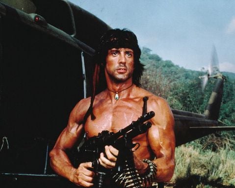 Rambo stands next to a helicopter and is holding a machine gun.