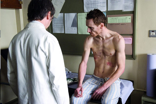 A gaunt, shirtless Michael Fassbender sits on an examination table with a doctor next to hime