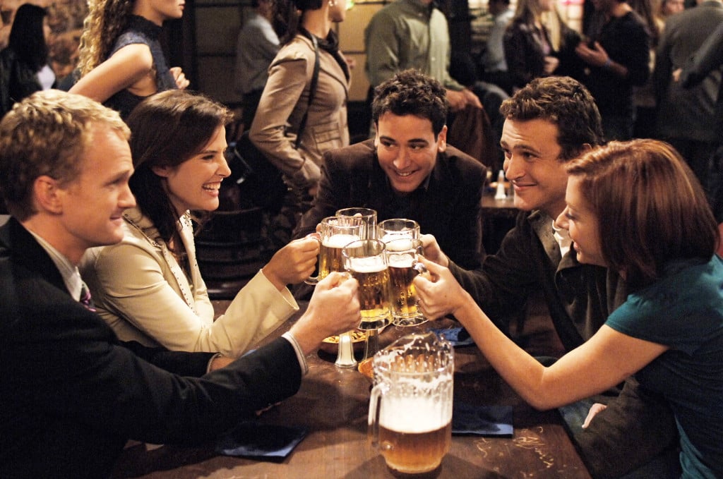 The cast of How I Met Your Mother smiling in a bar clinking their beer glasses together The cast of How I Met Your Mother smiling in a bar clinking their beer glasses together
