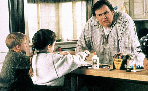Uncle Buck leans on a counter in front of two kids playing with crayons