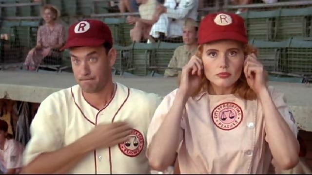Tom Hanks holds his hand over his heart while wearing a baseball uniform and standing next to Geena Davis in A League of Their Own