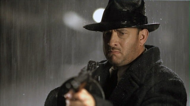 Tom Hanks holds up a shotgun while standing in the rain in a coat and fedora in Road to Perdition