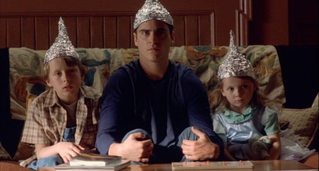Joaquin Phoenix sits next to two kids on a couch in foil hats in a scene from Signs