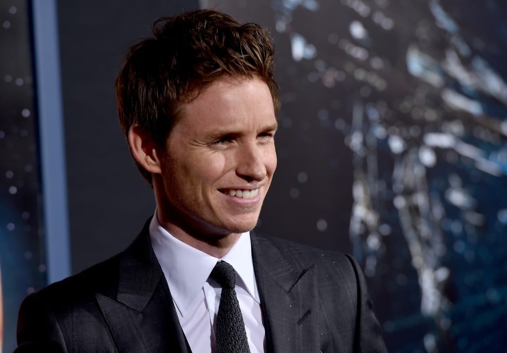 Eddie Redmayne smiles while wearing a dark suit and black tie. Eddie Redmayne smiles while wearing a dark suit and black tie.
