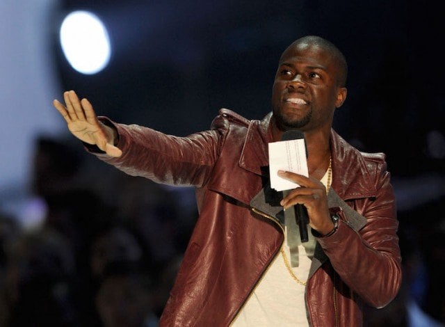 Kevin Hart extends one arm forward as he holds a microphone with his hand.
