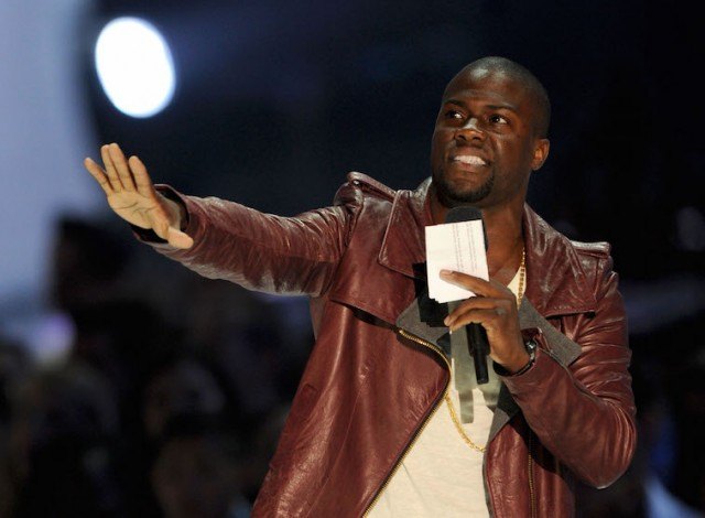 Kevin Hart extends one arm forward as he holds a microphone with his hand. Kevin Hart extends one arm forward as he holds a microphone with his hand.