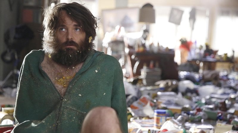 A bearded man sits on a messy floor in a green hoodie