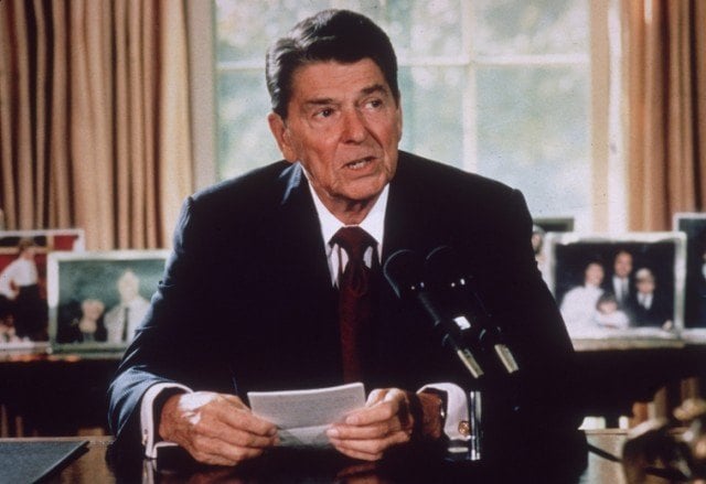 President Reagan speaks into a microphone on his desk while holding note cards. 