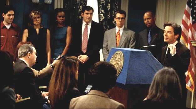 Michael J. Fox stands at a podium in front of a crowded room in Spin City