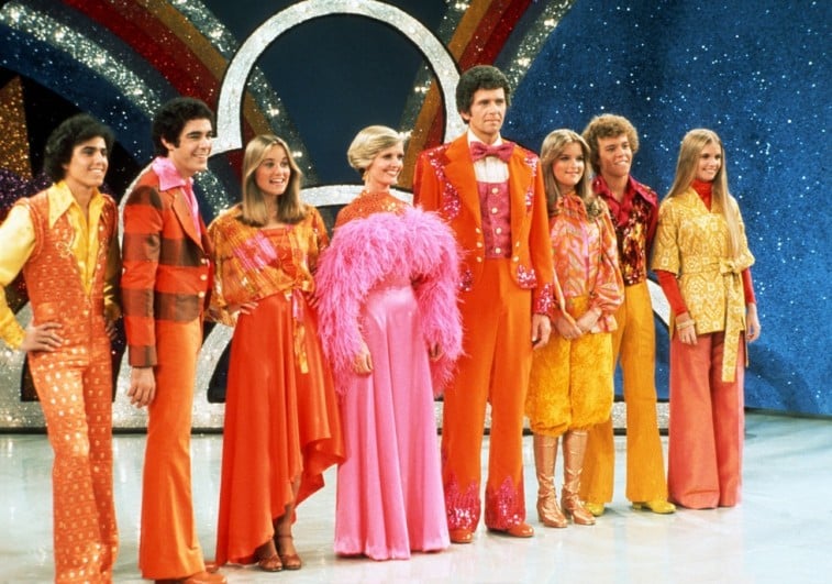 El reparto de la Tribu de los Brady se sitúa en el escenario en una fila mientras lleva coloridos trajes en La hora de los Brady