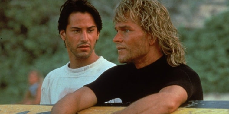 Keanu Reeves looks at Patrick Swayze as they sit on a beach in Point Break.