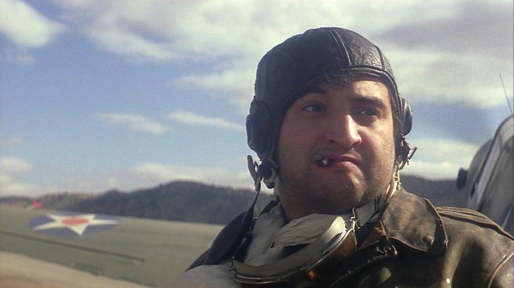 John Belushi is dressed as a pilot with a cigar in his mouth.